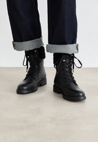 Stivali neri in pelle fino alla caviglia con punta arrotondata, superficie texturizzata e accessori in oro. Abbinati a jeans in denim scuri arrotolati. Suola piatta.