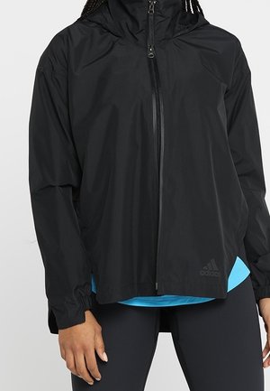 Personne portant une veste noire zippée Adidas avec une capuche, un t-shirt bleu en dessous et un legging noir, debout les bras détendus le long du corps.