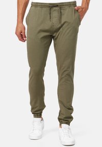 Muž v olivově zelených teplákových kalhotách se stahovací šňůrkou v pase a elastickými manžetami, obutý do bílých tenisek, stojící před jednobarevným pozadím.