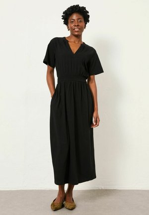 Schwarzes Maxikleid mit kurzen Ärmeln, V-Ausschnitt, gesmokter Taille und fließender Silhouette. Kombiniert mit braunen Slip-On-Schuhen vor hellem Hintergrund.