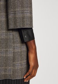 Gros plan d'une main à la peau foncée à côté d'une manche de veste à carreaux marron avec un poignet de chemise noir comportant deux boutons noirs.