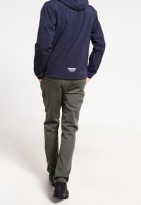 Veste à capuche bleu marine en tissu texturé, pantalons gris et chaussures noires. La veste présente un logo blanc et des détails de coutures au dos.