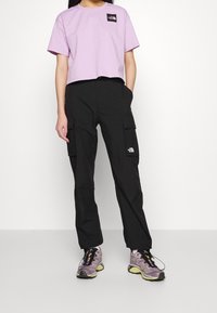 Lila kortärmad t-shirt med svart logotyp, kombinerad med svarta cargo-byxor med sidofickor. Grå sneakers med gula detaljer.