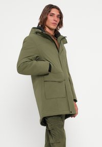 Olijfgroene parka jas met capuchon, voorzien van een ritsluiting, grote voorzakken en een licht oversized pasvorm met een zachte textuur.