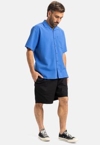 Camisa de manga corta azul con tela texturizada, cierre de botones y un bolsillo en el pecho, combinada con pantalones cortos negros y zapatillas negras.