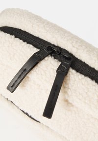 Fleecová taška s krémovým vonkajším povrchom a čiernym zipsom. Disponuje dvojitými čiernymi ťahadlami zipsu. Mäkká textura s hladkým vnútorným podšívaním.