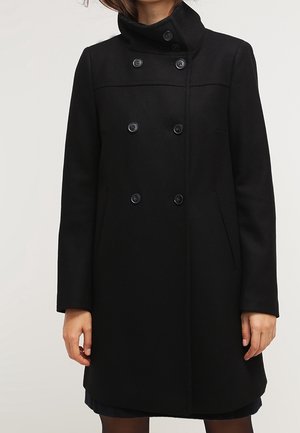 Sort dobbeltradet frakke med seks knapper, høj krave, lange ærmer og sidelommer båret af en model med hænderne afslappet langs siderne.