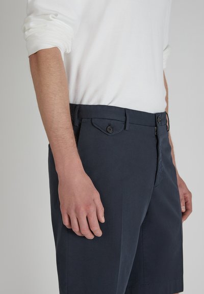 Pantaloni corti blu navy realizzati in un tessuto leggero, dotati di tasche laterali, una patta con bottone e una zip. Indossati con una camicia bianca a maniche lunghe.