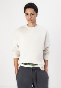 Pulover alb din tricot cu guler rotund și umeri căzuți, purtat împreună cu pantaloni de trening gri închis, cu un tiv elastic și un model în carouri verzi.