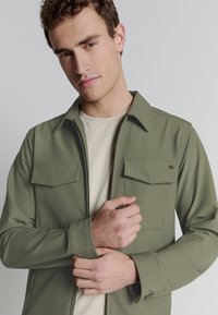 Olijfgroene lichte jas met twee borstzakken, een overhemdkraag en lange mouwen, gedragen over een beige ronde hals shirt.