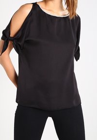 Black, short-sleeve blouse in smooth, lightweight fabric. Features cut-out shoulders, knotted sleeves, and a pearl-accented neckline.