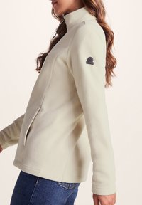 Light beige fleece jacket with a high collar, side zipper pockets, and a logo patch on the left sleeve. Smooth texture, casual design.