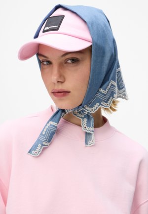 Femme portant une casquette de baseball rose avec un patch logo noir et une écharpe bleue brodée nouée sous le menton par-dessus la casquette.