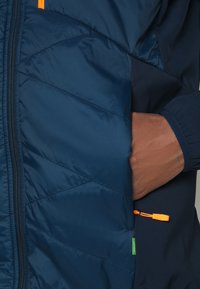 Marinblå puffjacka med ett kviltat mönster, dragkedjestängning och orange dragkedjehandtag; inkluderar sidofickor och en texturerad insida.