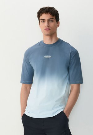 Jeune homme aux cheveux bouclés, debout face à l’avant, portant un t-shirt à manches courtes dégradé bleu-blanc avec le texte « Copenhagen » sur la poitrine.