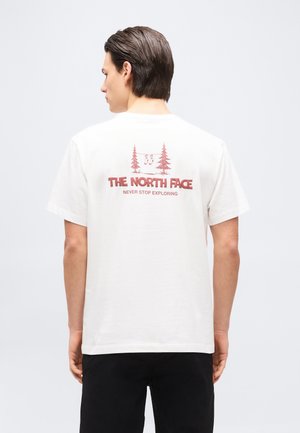 Hombre con camiseta blanca de manga corta con logo rojo de "The North Face" y texto "Never Stop Exploring", de espaldas sobre fondo liso.
