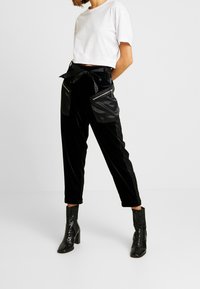 Femme debout portant un t-shirt blanc court, un pantalon en velours noir avec de grandes poches zippées, une ceinture nouée et des bottes noires à talons hauts.