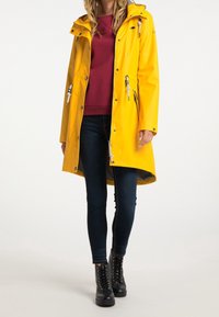 Žlutý nepromokavý plášť s kapucí, se stříbrnými zipy, nastavitelnými šňůrkami a volným střihem. Nesený přes vínový svetr a tmavé džíny.