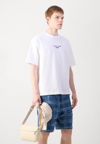 Camiseta blanca de algodón con el logo negro "Calvin Klein Jeans", combinada con shorts a cuadros azules. Los accesorios incluyen una bolsa cruzada beige y una gorra.