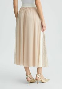 Gonna in tulle di un beige pallido con vita elasticizzata, caratterizzata da un pannello laterale con un motivo. Abbinata a scarpe con tacco metalliche dorate.