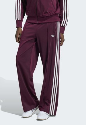 Persona che indossa pantaloni sportivi Adidas color vino con tre strisce bianche ai lati e scarpe da ginnastica bianche, in piedi su uno sfondo chiaro.