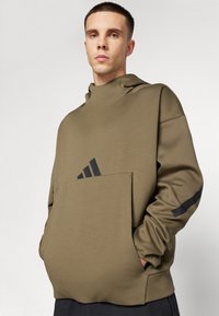 Hoodie khaki con colletto alto e tasche laterali. Presenta dettagli neri e un logo sul davanti. Materiale morbido e leggero.