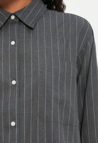 Camicia grigia con bottoni e strisce verticali bianche, caratterizzata da un colletto classico e una tasca sul petto sinistro. Realizzata in un tessuto testurizzato.