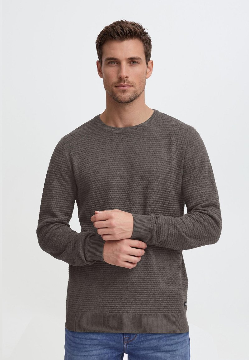 Mand med kort brunt hår iført en tekstureret mørkegrå sweater og blå jeans, stående med hænderne foldet foran.