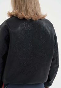 Černá kožená bunda s embosovanými květinovými a hadími vzory na zadní straně, uvolněný střih a hladká textura.
