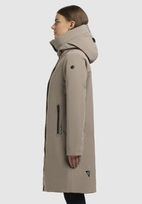Lange beige jas met een hoge kraag en grote capuchon, voorzien van twee ritssluiting zijzakken en een driehoekig logo nabij de zoom. Glad van textuur.