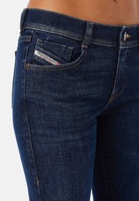 Jeans ajustados de mezclilla oscura con una textura suave. Presenta costuras en contraste y una pequeña etiqueta cosida en el bolsillo delantero. Diseño clásico de cinco bolsillos.