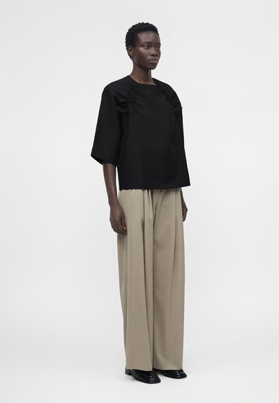 Γυναίκα όρθια, φοράει μαύρο χαλαρό τοπ με βολάν και μπεζ παντελόνι με φαρδιά πόδια, μαύρα παπούτσια, μπροστά σε απλό φωτεινό φόντο.