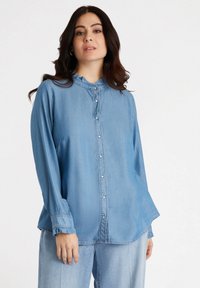 Camisa de denim azul claro con botones en la parte delantera, cuello con volantes, mangas largas y un corte relajado. Presenta puños fruncidos y un dobladillo curvado.