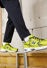 Personne portant des baskets New Balance jaune vif et un pantalon sombre à fines rayures marchant sur une plateforme en bois contre un mur en briques blanches.