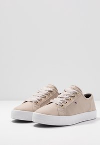 Beige Sneaker aus Canvas mit niedrigem Schnitt, weißen Gummisohlen, weißen und grauen gestreiften Schnürsenkeln, goldfarbenen Ösen und einem kleinen Logo in Form einer roten, weißen und blauen Flagge.