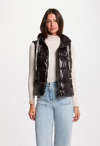 Gilet nero imbottito con cappuccio, finitura lucida, due tasche laterali e cordini bianchi. Presenta una zip frontale ed è indossato su un maglione crema.