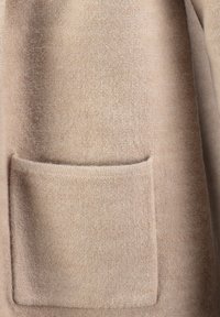 Tessuto di lana beige con una texture morbida, dotato di una tasca rettangolare a toppa. Design semplice e senza cuciture, privo di hardware aggiuntivo.