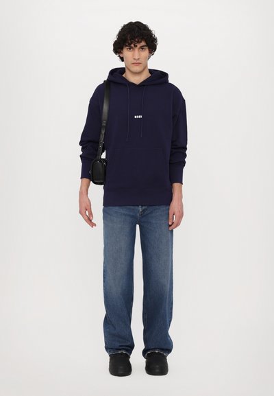 Navy hoodie with front pocket and drawstring, paired with light blue wide-leg jeans and black shoes. Small white logo on the hoodie.