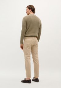 Pantalones de algodón beige y un suéter de canalé verde oliva, con un diseño clásico y un tejido texturizado. El modelo lleva zapatos oscuros.