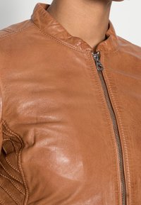 Bruine leren jas met een gladde textuur, een staande kraag en een zilveren ritsdetail aan de voorkant. Verstevigde stiksels op de mouwen.