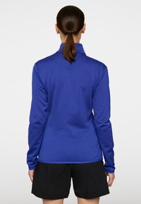 Polo a maniche lunghe blu, tessuto liscio, colletto alto, design aderente, abbinato a pantaloni corti neri. La vista posteriore mostra i dettagli delle maniche e dell'orlo.