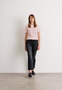 Kvinne som går innendørs iført en lys rosa kortermet topp, svarte høytalende rette jeans og svarte loafers foran en hvit vegg.