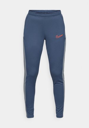 Námořnické sportovní kalhoty s pasem na tkaničku, šedými bočními panely a oranžovým logem Nike na levé noze. Hladká, pružná látka.