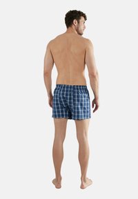 Boxer da uomo a quadri blu e bianchi con vita elastica, mostrati da dietro su un modello maschile con la pelle nuda, in piedi su piedi nudi.