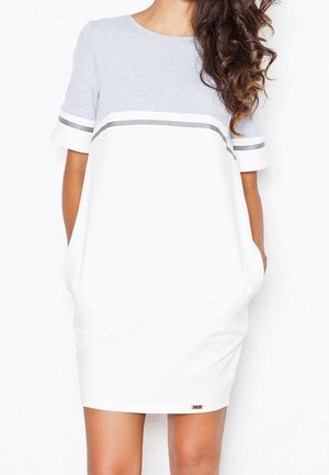 Mujer con un vestido de manga corta y diseño de bloques de color, con la parte superior gris claro y la falda blanca con bolsillos, de pie frente a un fondo blanco.