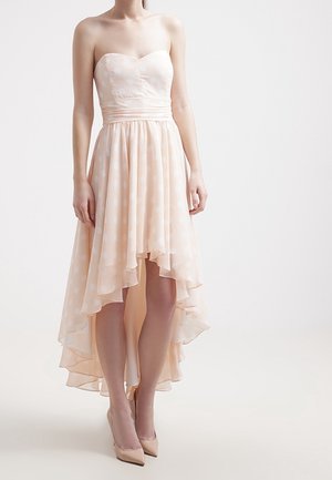 Trägerloses, blassrosa High-Low-Kleid mit dezenten Polka-Dots, tailliertem Oberteil, fließendem Rock, getragen mit nude-farbenen spitzen High Heels an einem stehenden Model.