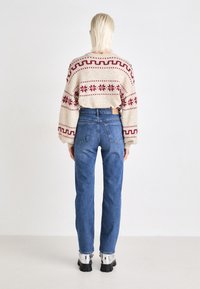 Μπεζ πουλόβ�ερ με κόκκινες πλέκες, συνδυασμένο με μπλε τζιν ψηλής μέσης. Τα παπούτσια έχουν ασημί λεπτομέρειες και χοντρές μαύρες σόλες.