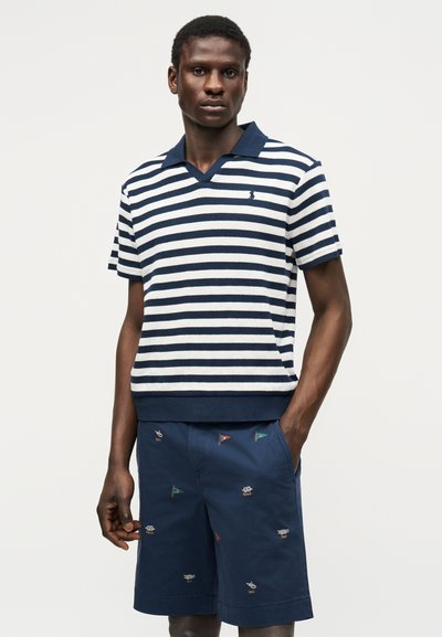Άνδρας που φοράει μπλε-άσπρο polo με κοντά μανίκια και οριζόντιες ρίγες, καθώς και σκούρο μπλε σορτς με μικρές κεντημένες σημαίες, στέκεται με το ένα χέρι στην τσέπη.