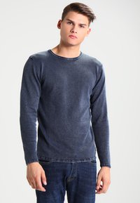Maglietta a maniche lunghe blu navy con collo rotondo realizzata in tessuto morbido, con una texture leggermente sbiadita e dettagli di cucitura visibili. Indossata con jeans scuri.