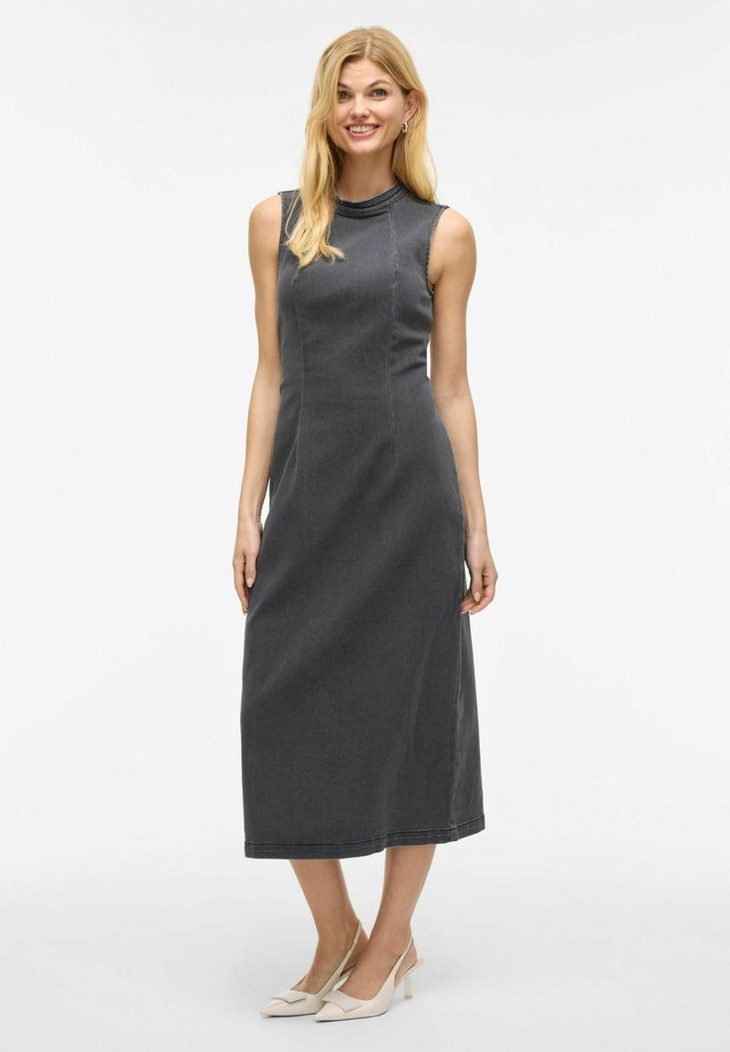 Grå denim midi-klänning med hög halsringning, ärmlös design, figurnära silhuett och sömnadsdetaljer, i kombination med ljusa klackar.